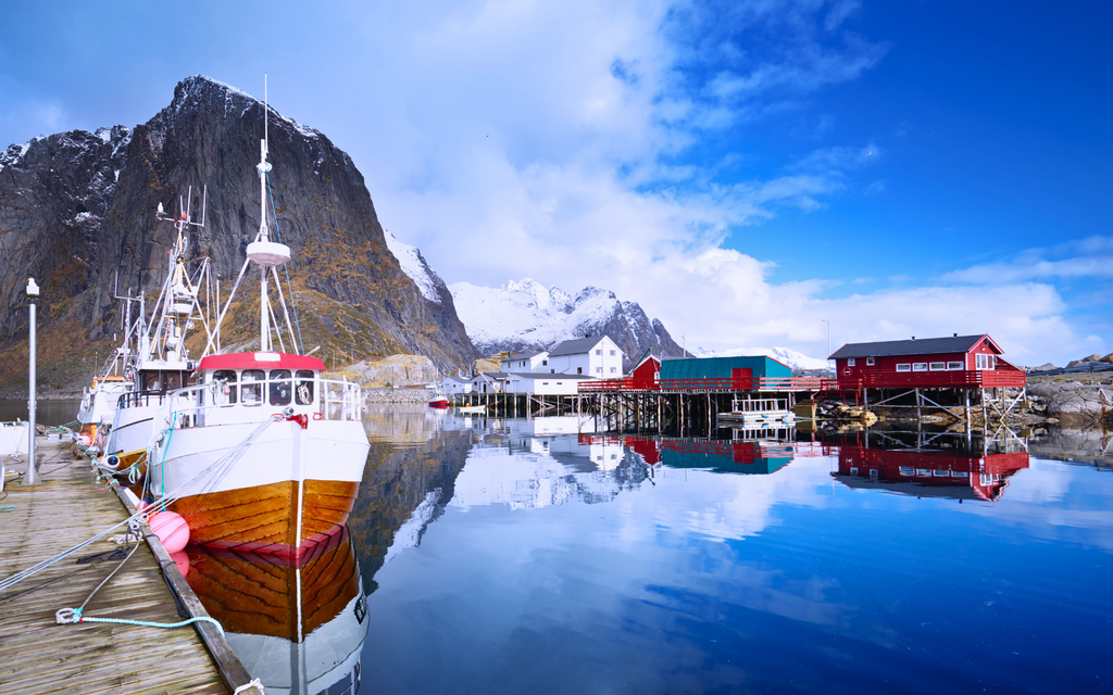 A scenic view of a Norwegian fishing village with traditional fishing boats docked in clear waters, surrounded by snow-capped mountains and rustic buildings, representing sustainable fishing practices.